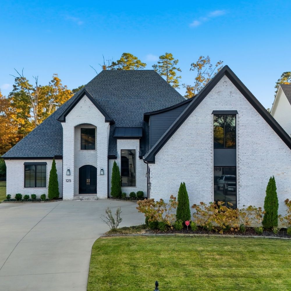 Modern two-story house with white brick exterior, black trim, large windows, black front door, neatly landscaped yard, curved driveway, and clear blue sky in the background.