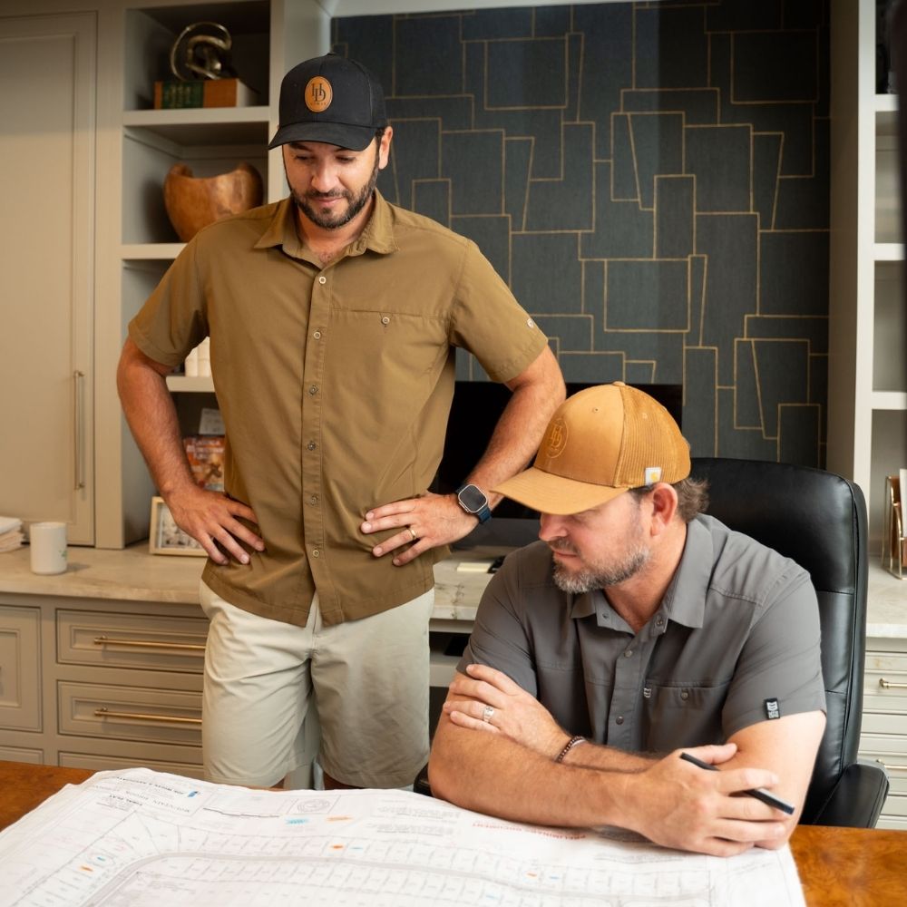 Two men wearing casual shirts and hats are looking at plans or blueprints on a desk in an office. One is seated with a pen in hand, while the other stands beside him, appearing to discuss the documents.