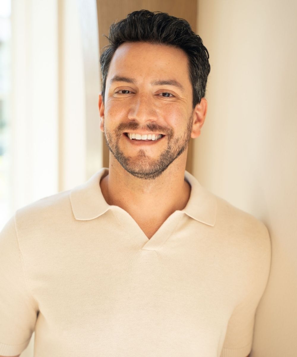 A man with short dark hair and a trimmed beard smiles while leaning against a light-colored wall, wearing a beige collared shirt in soft natural light.