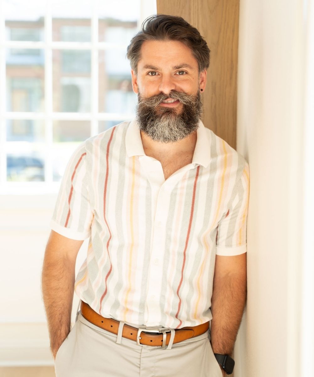 A smiling man with a full beard and mustache, wearing a light-colored, short-sleeve, striped shirt and beige pants, stands indoors by a sunlit window, leaning casually against a wall.