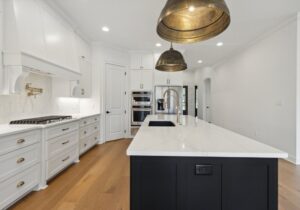Modern kitchen with white cabinets, wood flooring, a large white marble island with a black base, gold pendant lights, built-in ovens, a gas cooktop, and brass fixtures. The space is bright and spacious.