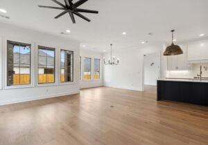 Open concept living and dining area with light wood floors, large windows, white walls, black ceiling fan, modern chandelier, and a kitchen with white cabinets and brass pendant lights.