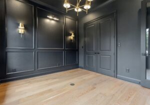 A modern, empty room with dark paneled walls, double doors, light wood flooring, two wall sconces, and a gold chandelier. The room appears clean and minimalist with no furniture.