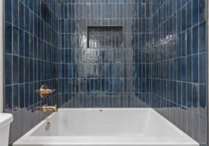 A modern bathtub with glossy, rectangular dark blue tiles on the surrounding walls, featuring a built-in niche and gold-colored faucet and shower controls on the left side.