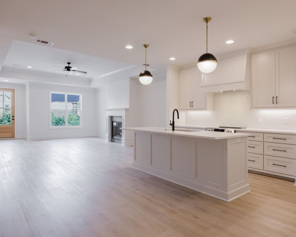 Spacious, modern open-plan kitchen and living area with light wood floors, white cabinetry, a large island with black faucet, pendant lights, fireplace, and large window overlooking greenery.