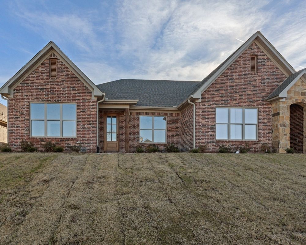 A single-story red brick house with large windows, a wooden front door, and a gabled roof sits on a grassy, slightly sloped lawn under a partly cloudy sky.