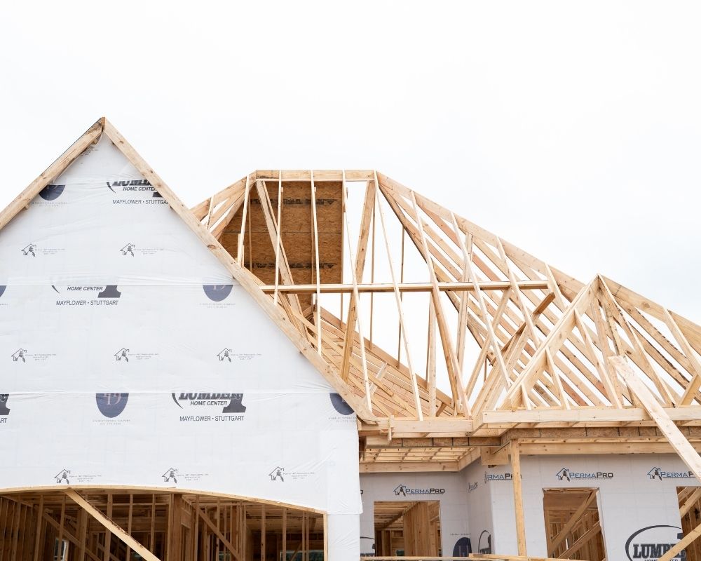 A house under construction with exposed wooden framing and roof trusses. The lower part of the house is wrapped in white protective sheeting. The sky above is overcast.