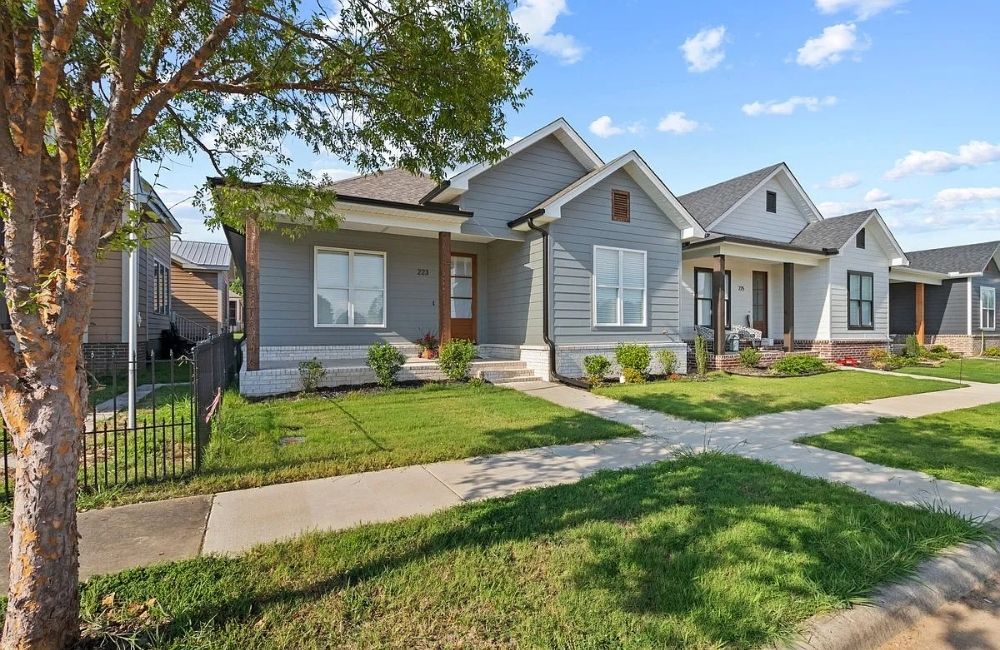 A modern, single-story gray house with white trim and a small front porch, surrounded by a well-kept lawn, shrubs, and a sidewalk under a partly cloudy sky. Neighboring houses are visible on both sides.