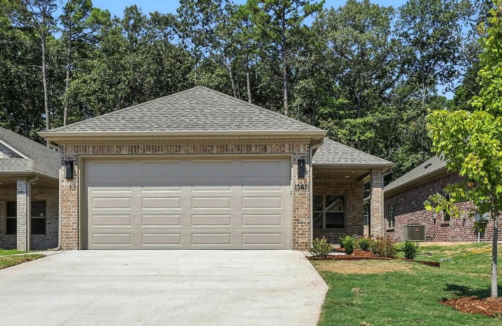 A single-story brick house with a two-car garage, light brown garage doors, and a sloped driveway. Green trees and shrubs surround the home, with a small lawn and a young tree in front.